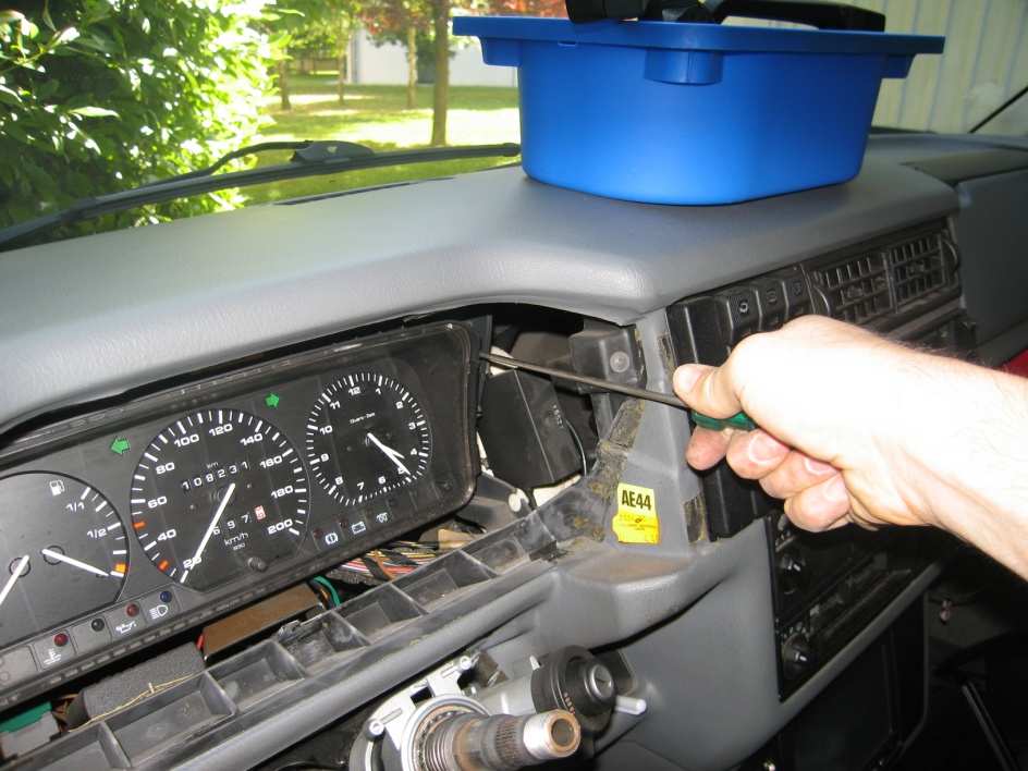A long screwdriver reaching past the side of the instrument cluster to the screws of the white clips