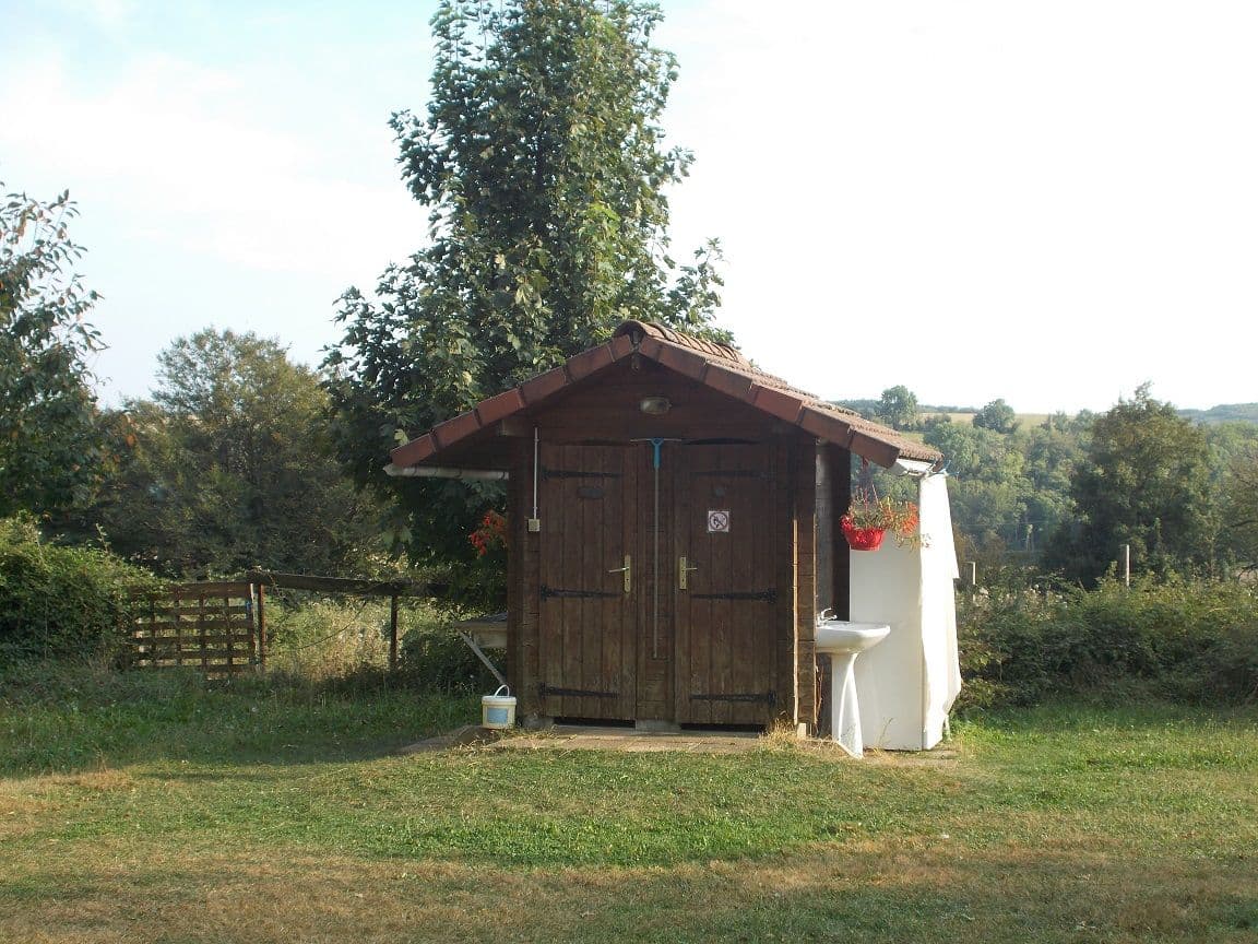The wooden hut with two doors housing the shower and toilet of the campsite, with a white basin on the side and the countryside behind