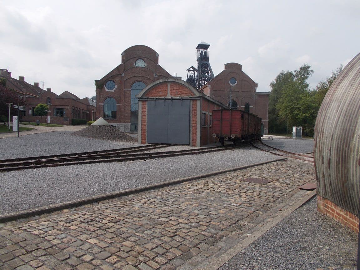 The brick buildings of the Le Bois du Cazier mine, with the tracks, a red wagon and cobblestones in the foreground