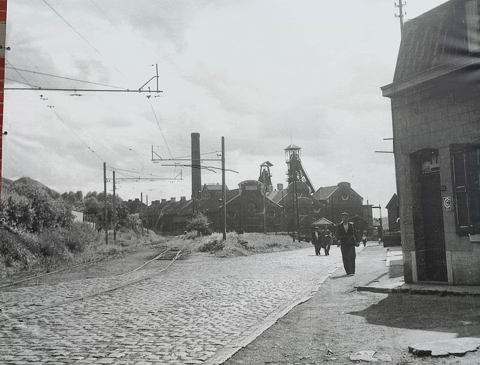 An old black-and-white photograph of the cobbled street with tracks, miners and the mine in the background