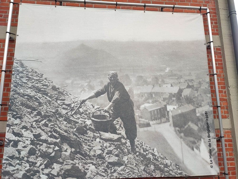 A large photographic panel of a woman collecting coal on the slag heap, with the village houses behind