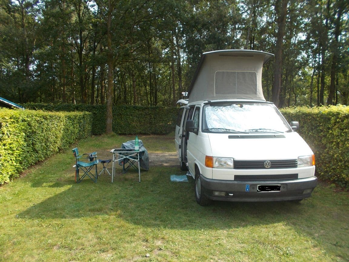 The T4 California with its roof raised on a pitch at the Zonhoven campsite, among hedges and trees, with chairs and a small table