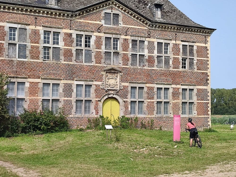 A red brick castle with a yellow door, a pink sign and a cyclist with her bike leaning in front