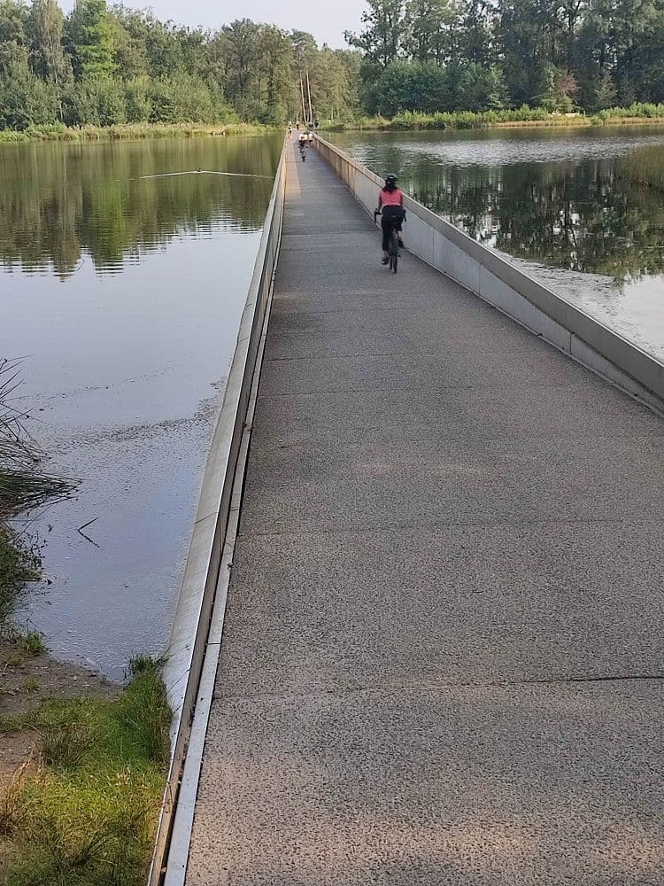 The cycle path crossing a small lake, with the water just centimetres from the edge and a few cyclists in the distance