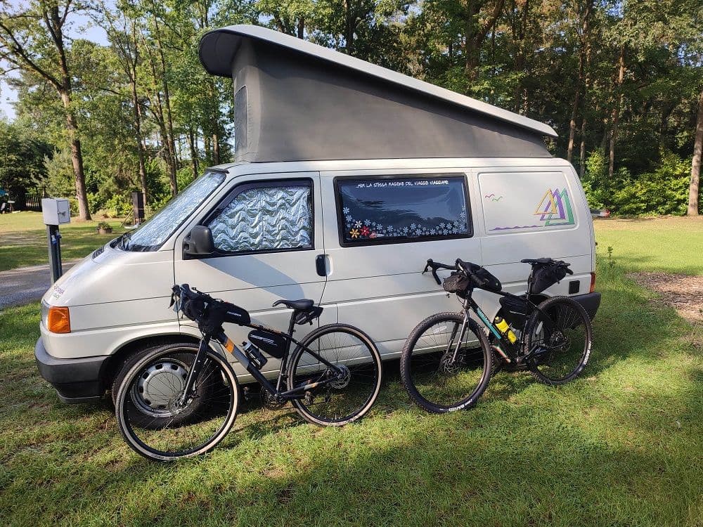The T4 California with its roof raised and two touring bikes with bags leaning against the van, among the trees of the campsite