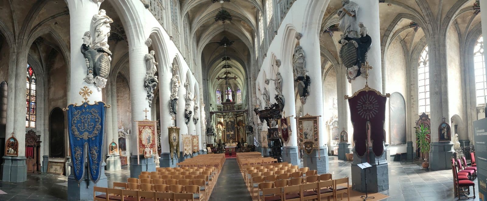 The nave of the church of St Sulpicius in Diest, with white columns decorated with statues and rows of wooden chairs