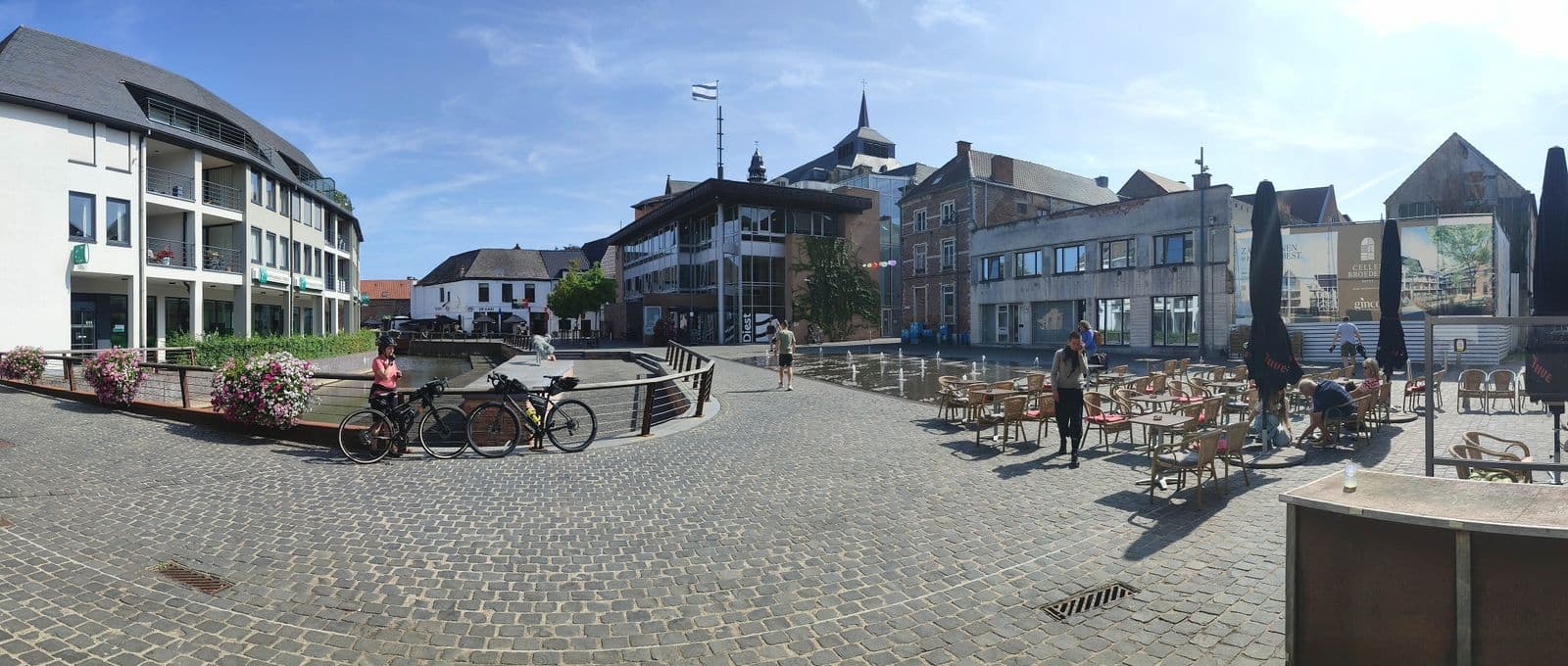 A cobbled square with café tables, a fountain and two bicycles leaning at the edge