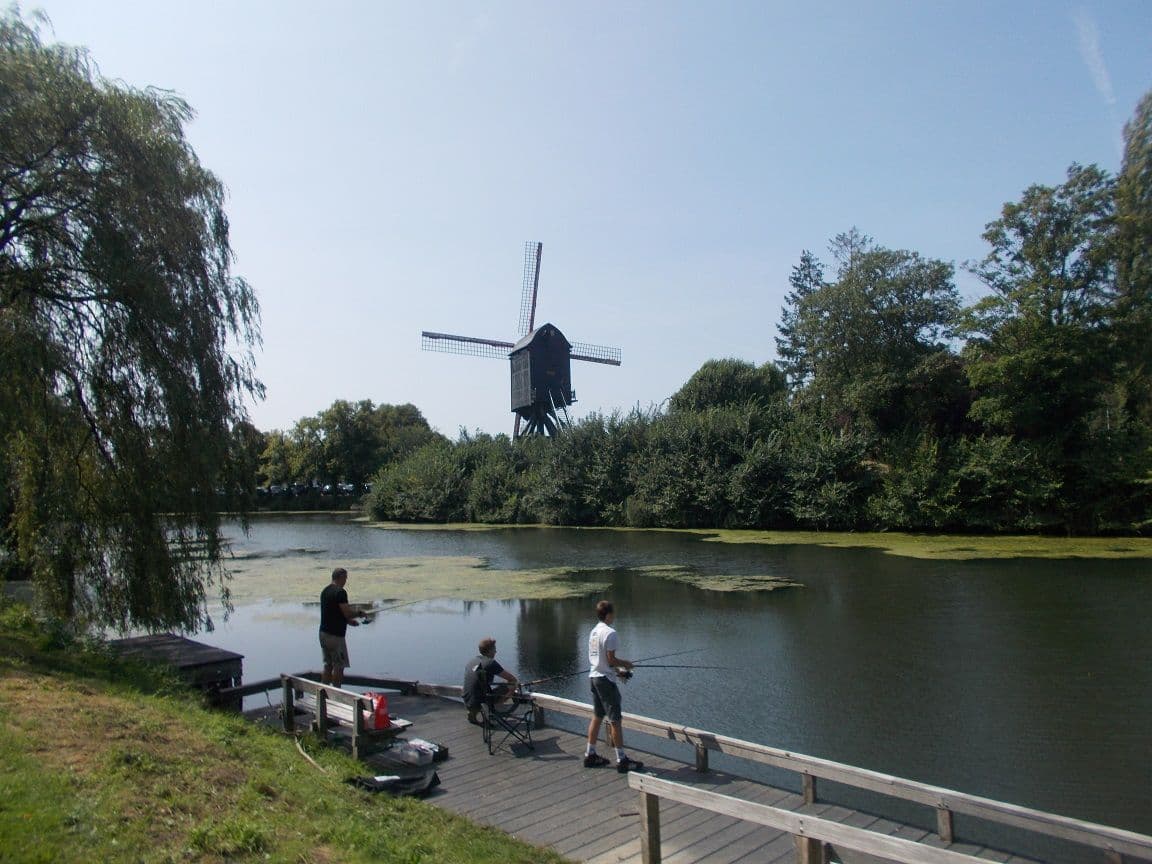 A windmill on the bank of a pond, with three anglers on a wooden jetty and a willow on the left